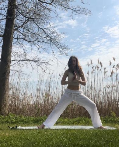 Woman in white tank top and pants in standing yoga pose, on grass with marsh grasses and tree in background with blue sky and clouds. 