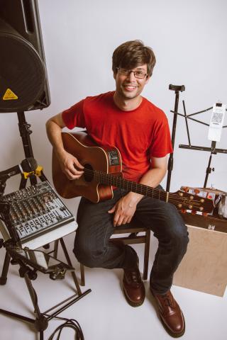 Musician seated wearing red shirt and black pants. Holding guitar and surrounded by other instruments.