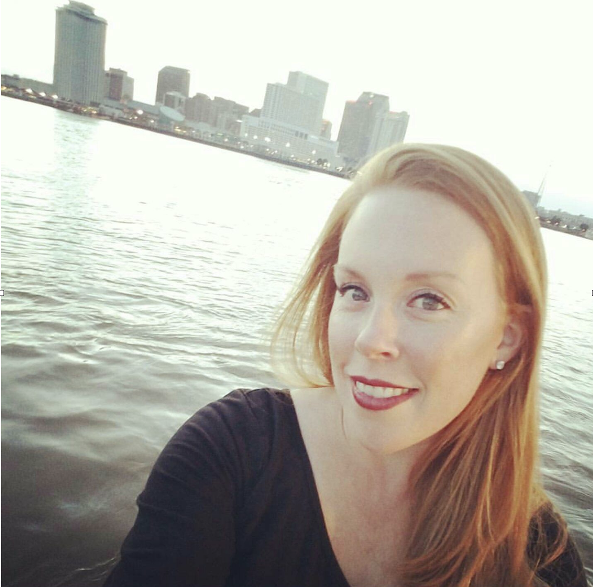 A photo of a woman with water and buildings behind her.